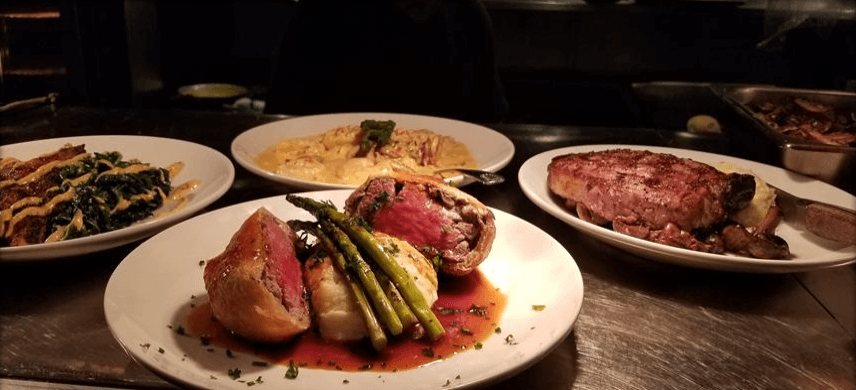 Kitchen pass shot of upscale entrées — a wrapped beef tenderloin with asparagus, a seared steak, pasta, and creamed spinach