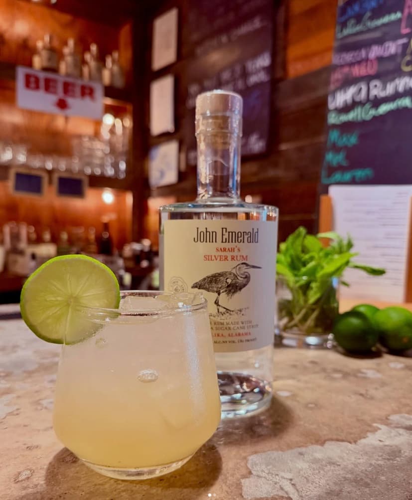 A lime-garnished cocktail in front of a bottle of John Emerald's Sarah's Silver Rum on the tasting-room bar