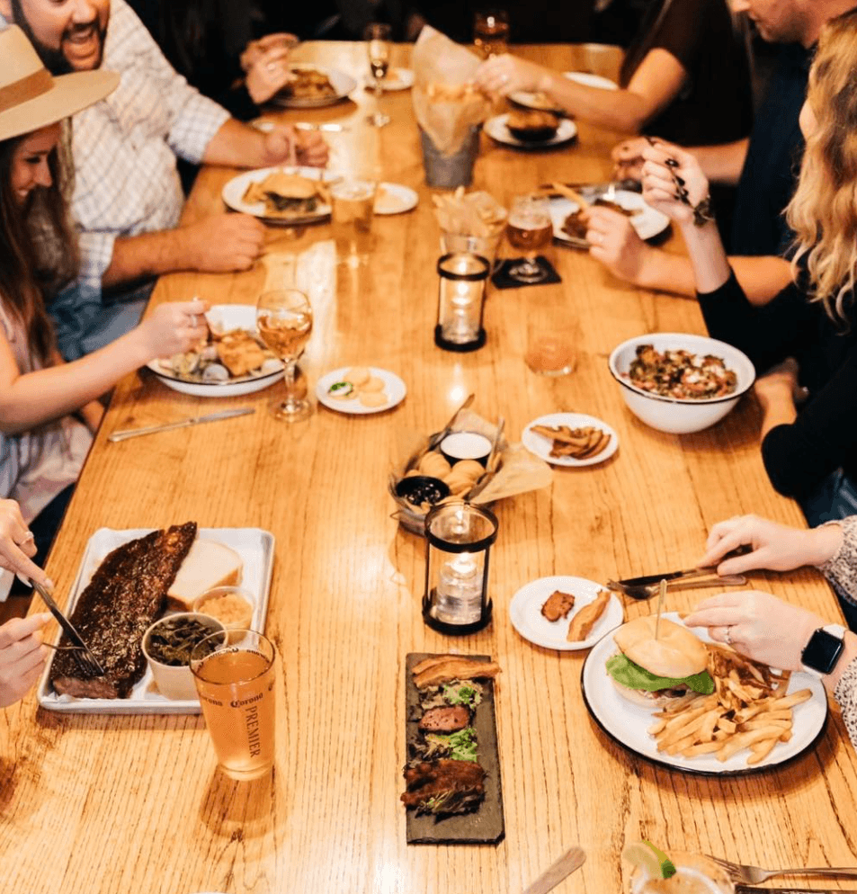 A group gathered around a long wood table loaded with ribs, burgers, fries, and drinks, lit by candle lanterns