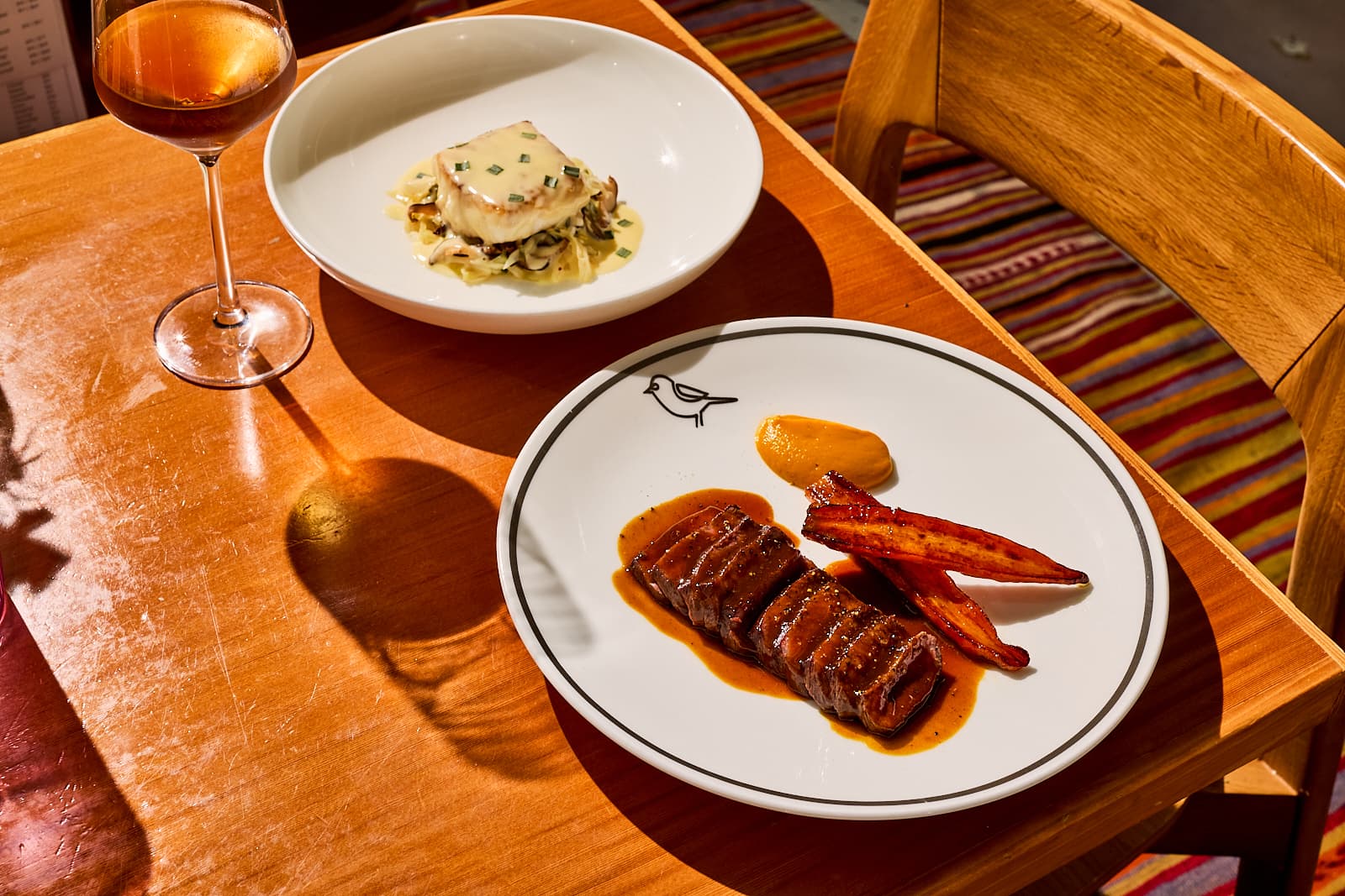 A seared duck breast with candied carrots on a branded Birdie's plate, alongside a cheese-topped dish over braised greens and a glass of rosé