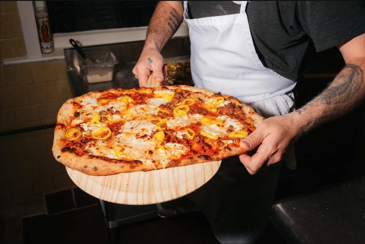 A cook at Little Donna's sliding a fresh tavern-style pizza with sausage and banana peppers off the peel