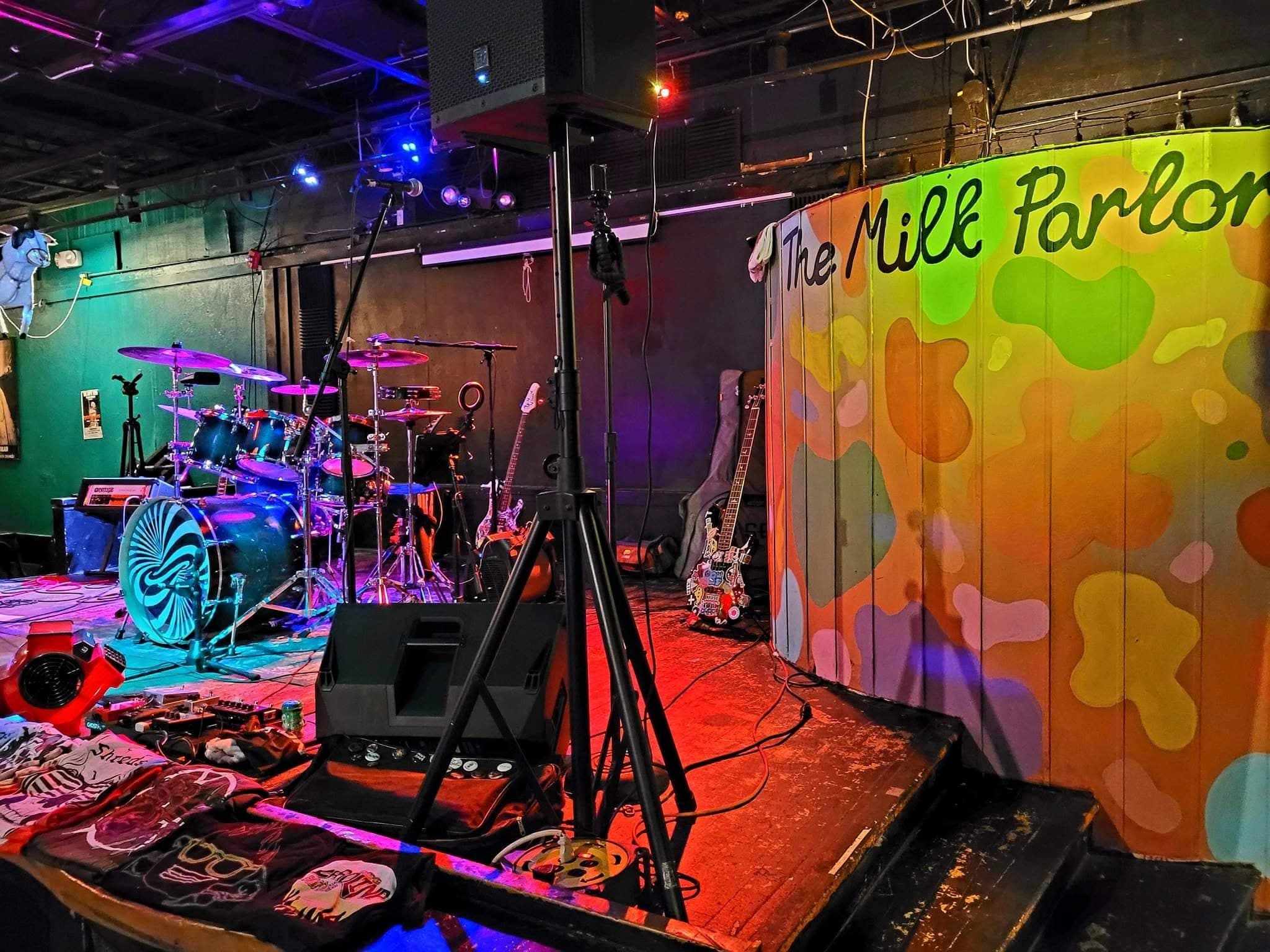 The Milk Parlor's stage set up with drums and guitars beside its hand-painted logo mural