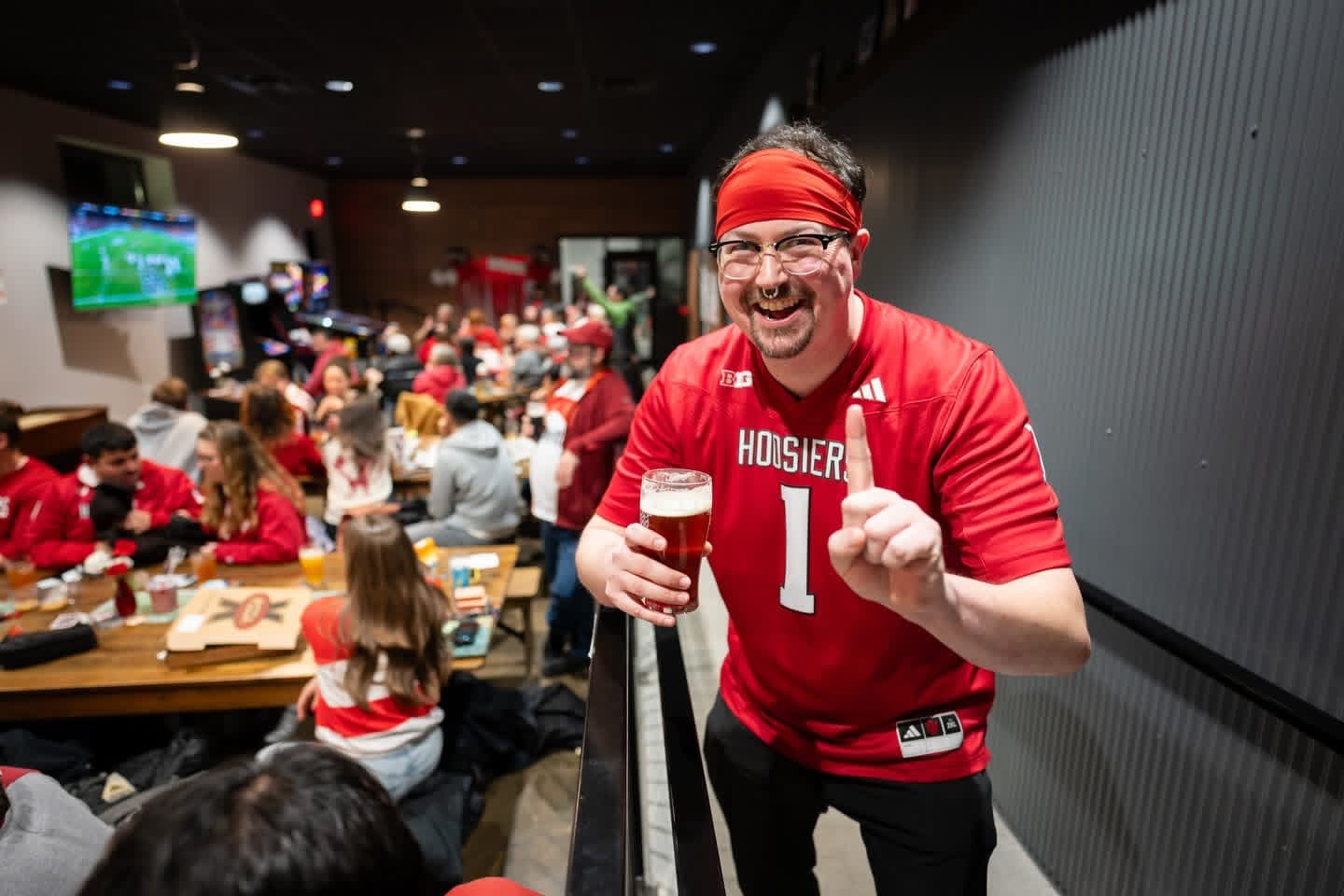 A crowded Hoosiers gameday scene inside the taproom, with a fan in a Hoosiers jersey holding a beer in the foreground