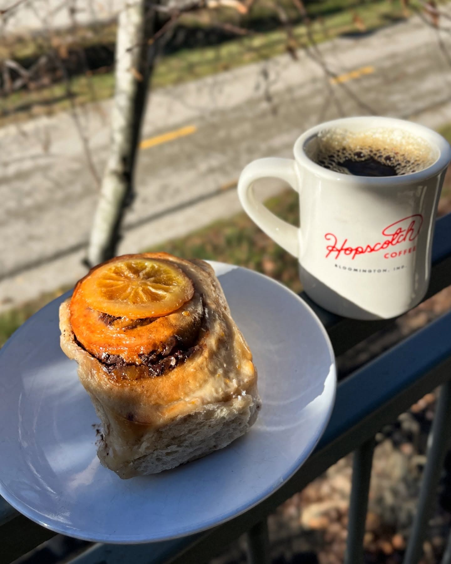 A branded Hopscotch Coffee mug next to a citrus pastry, sitting outdoors on a railing