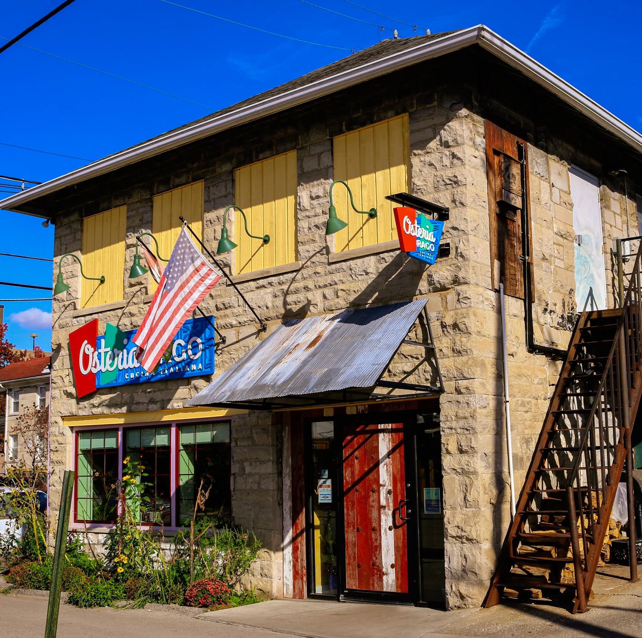 Sunlit exterior of Osteria Rago's limestone building with its red-and-blue signage over the front door