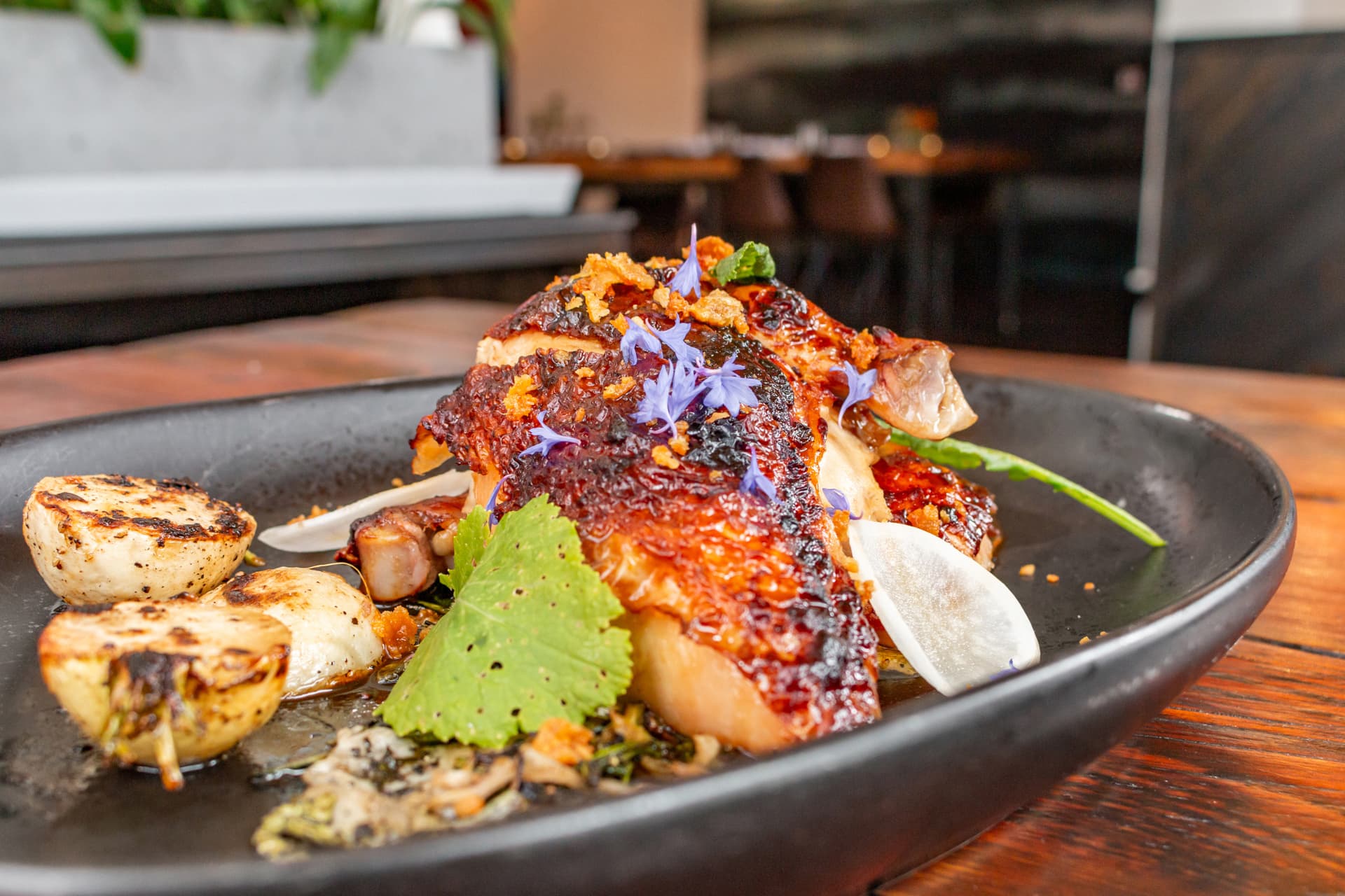 A glazed, charred plated dish topped with edible flowers next to grilled scallions at Blackbelly Market's dining room