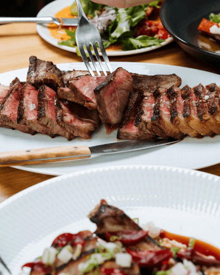 A sliced steak fanned out on a white plate at Corrida, with a knife alongside and salads in the background