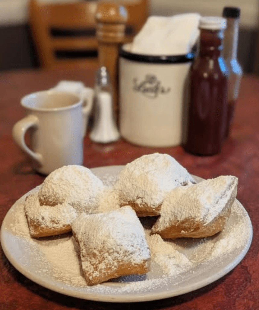 Four powdered-sugar beignets on a plate with a mug of coffee at Lucile's Creole Cafe