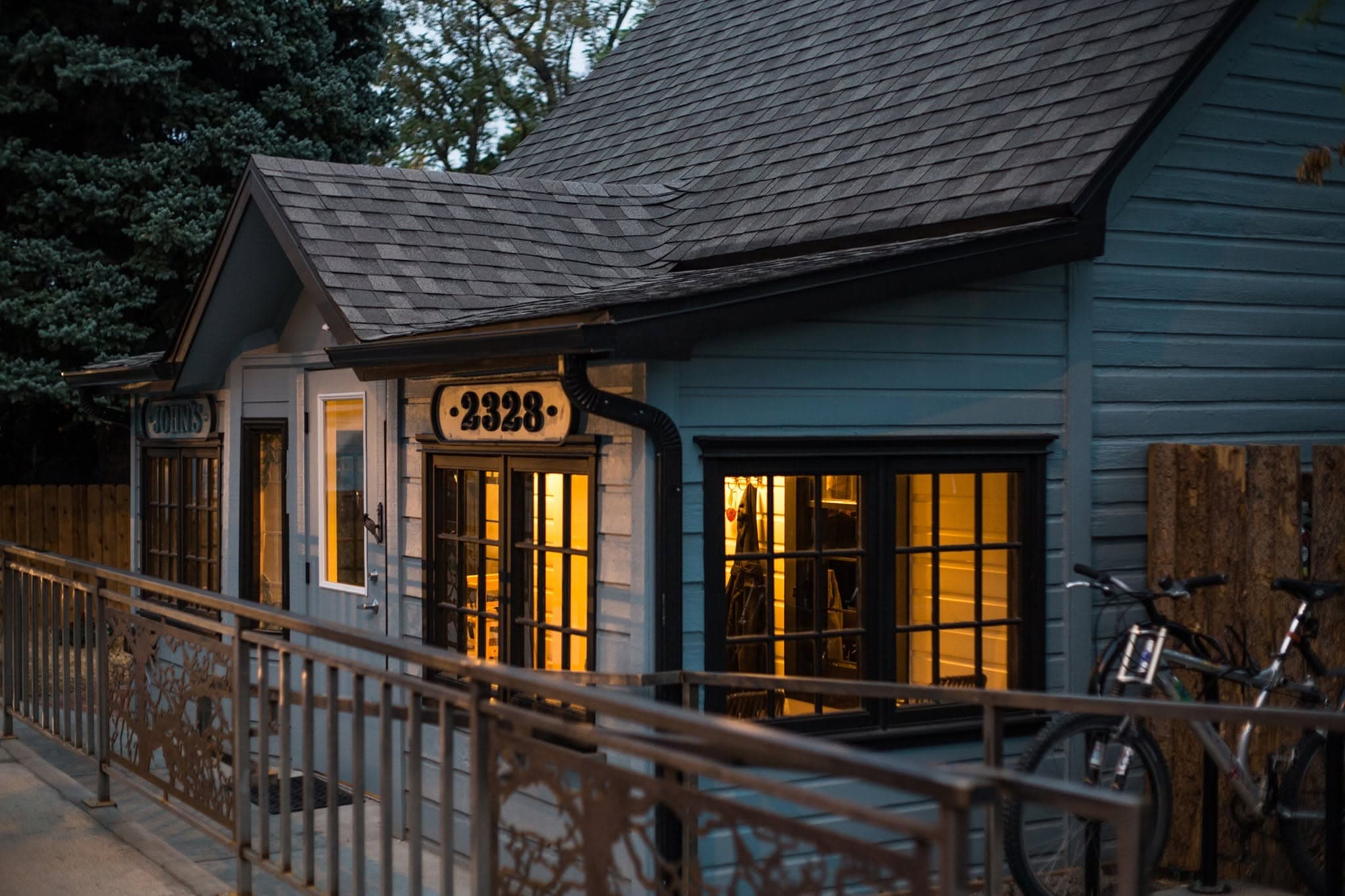 Twilight exterior of River and Woods' converted miner's-cabin building, warm light glowing through the windows and the old 'John's' sign still on the porch