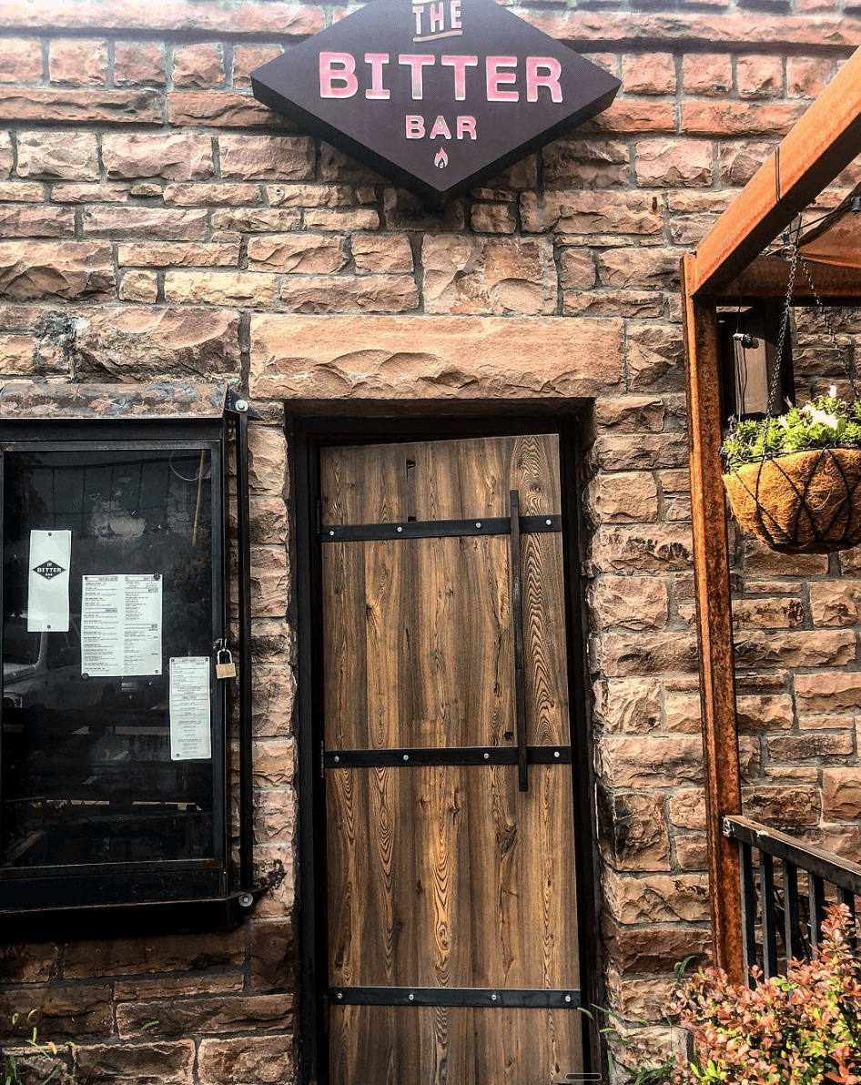 The Bitter Bar's stone-and-wood entrance with its diamond-shaped illuminated sign above the door and the posted menu beside it