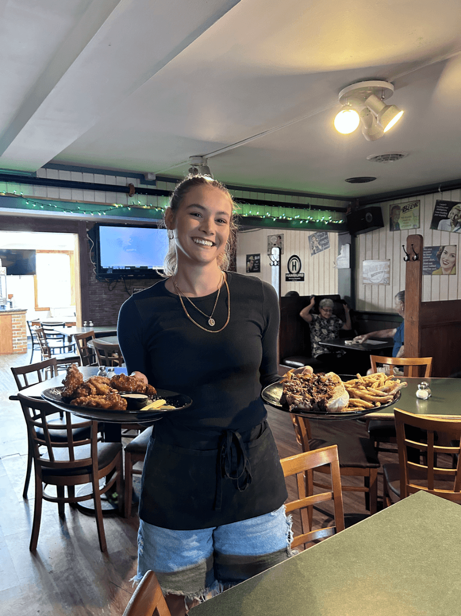 A smiling server at O'Neill's Stadium Inn holding a plate of wings and a loaded sandwich with fries