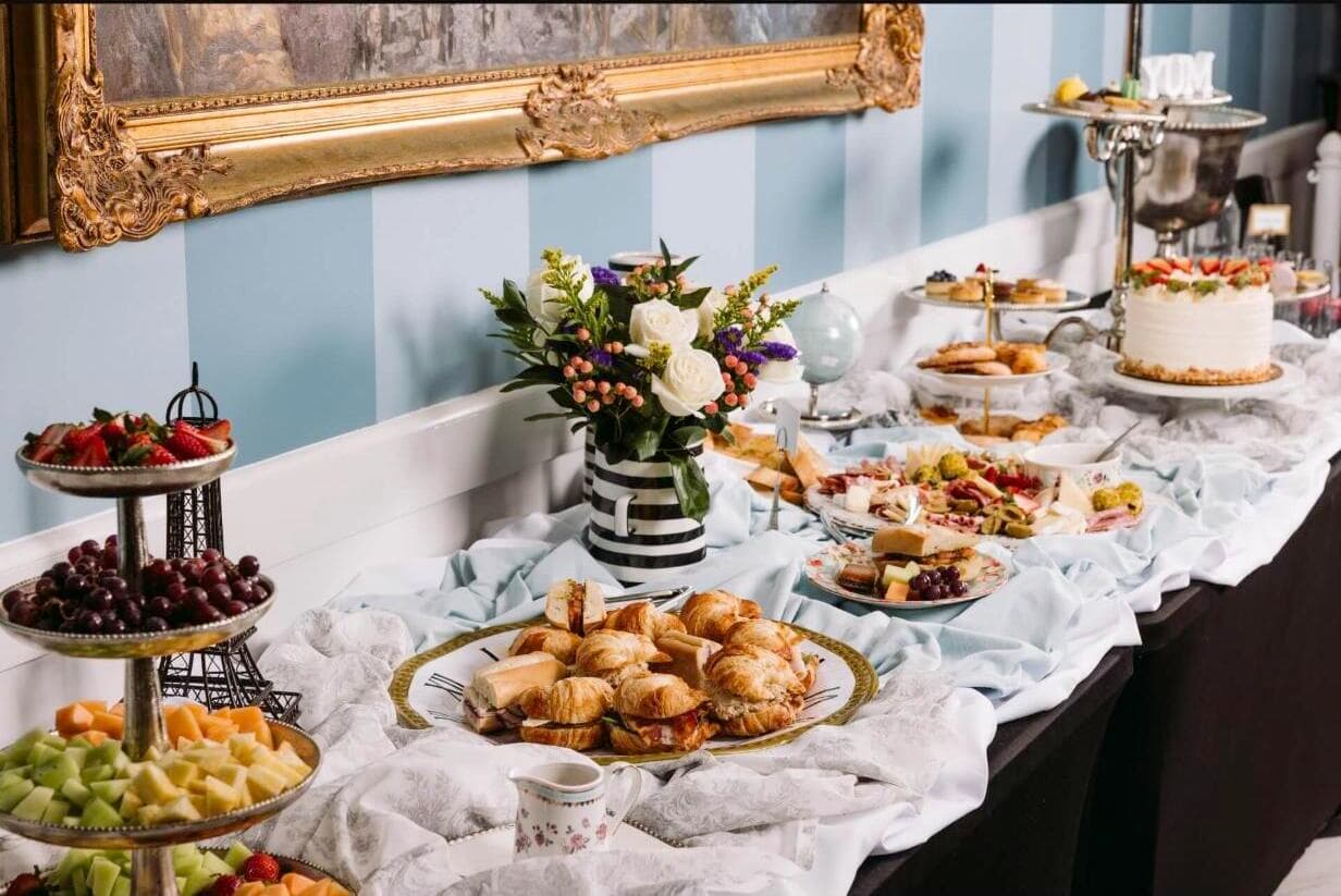 A long tea-party style spread at Amélie's — tiered trays of fruit, croissant sandwiches, a charcuterie board, pastries, and a frosted cake
