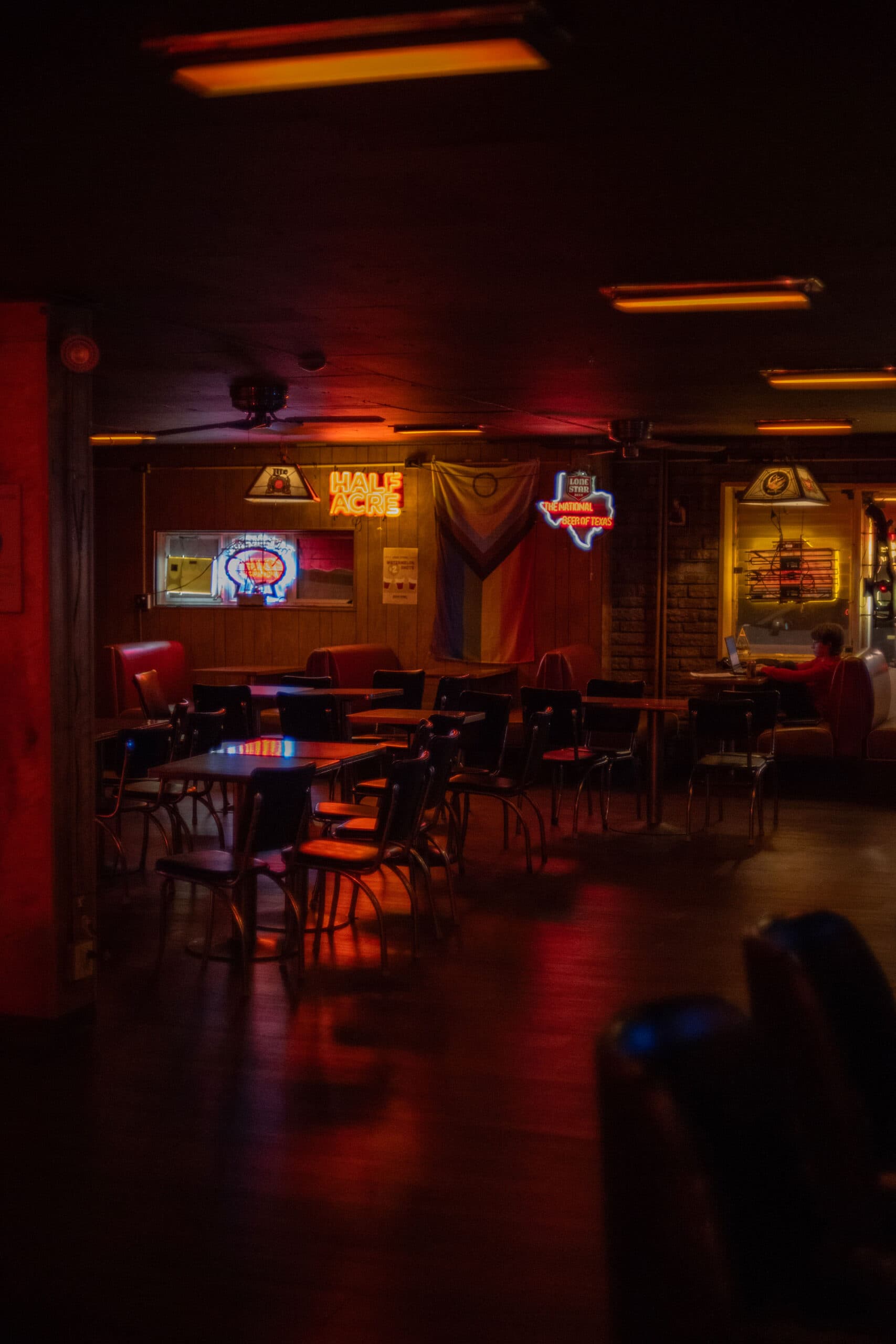 Empty honky-tonk dining room glowing with neon beer signs and a Pride flag hanging on the wood-paneled wall