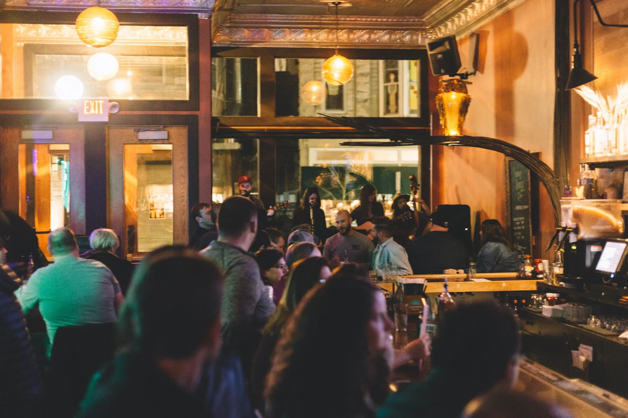 A packed crowd at the bar watching a band perform live under an ornate pressed-tin ceiling