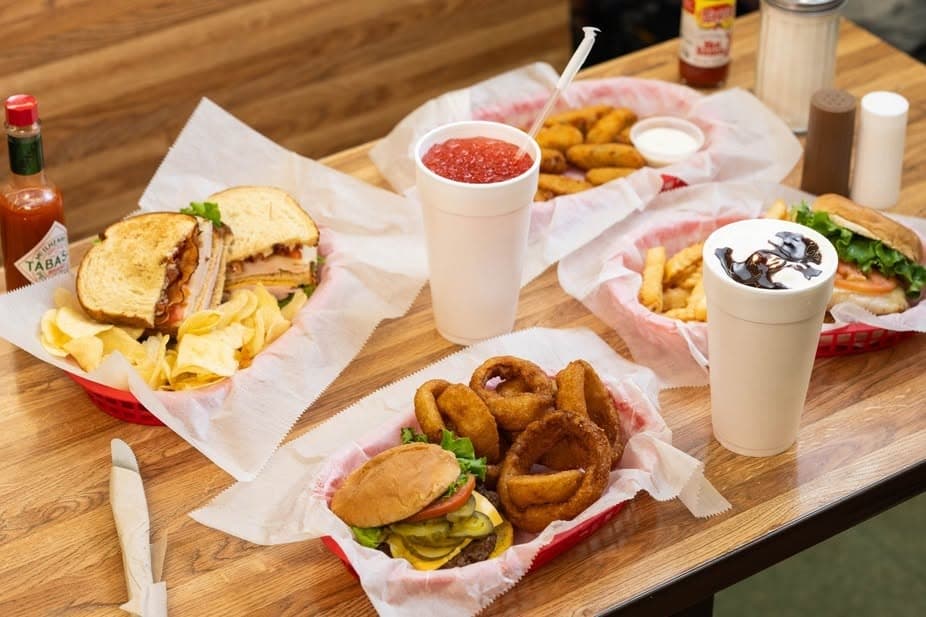 A diner table spread at Sutton's Drug Store with a club sandwich, onion rings, a burger and fries, and milkshakes