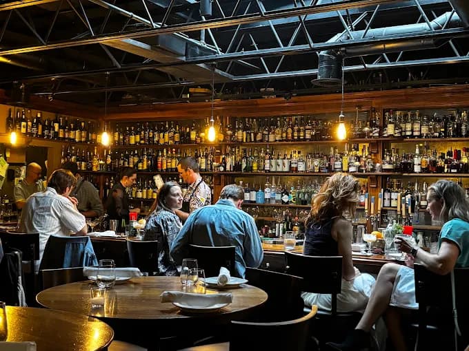 Guests seated at tables and the bar inside The Alley Light, backed by a wall of bottles beneath exposed-beam ceilings and warm hanging bulbs