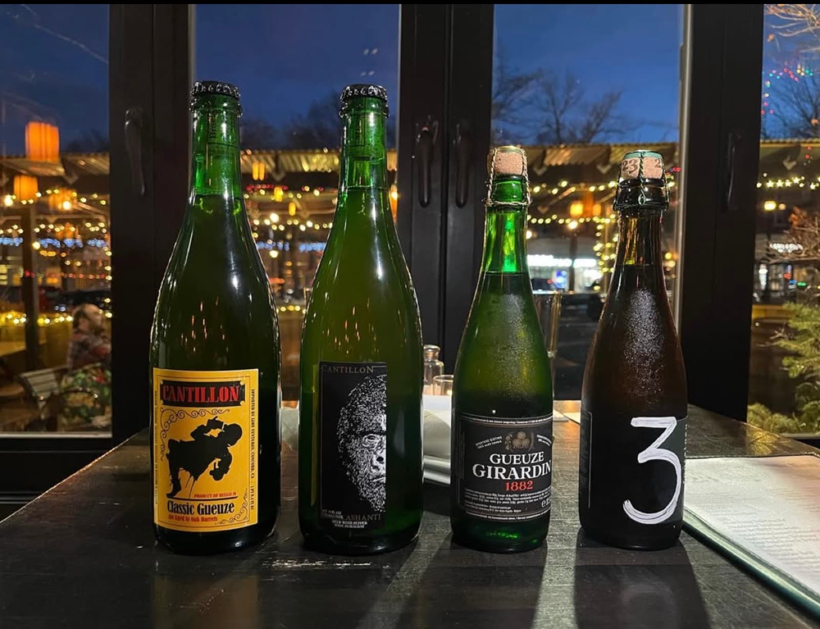 Four rare Belgian bottles — including two Cantillon labels — lined up on The Publick House's bar with string lights outside