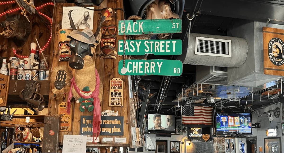 Eclectic dive-bar interior with a gas-masked deer mount, tiki masks, street signs, and TVs crowding the walls