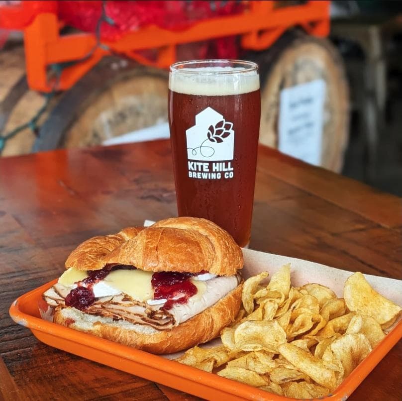 A branded Kite Hill Brewing Co. pint glass next to a croissant sandwich and chips on an orange tray
