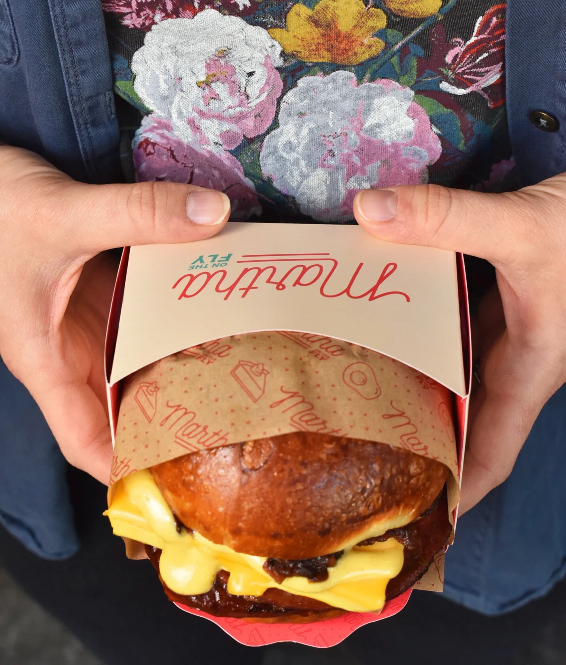 Two hands holding a branded Martha on the Fly box wrapped in butcher paper, opened to reveal a cheese-covered breakfast sandwich