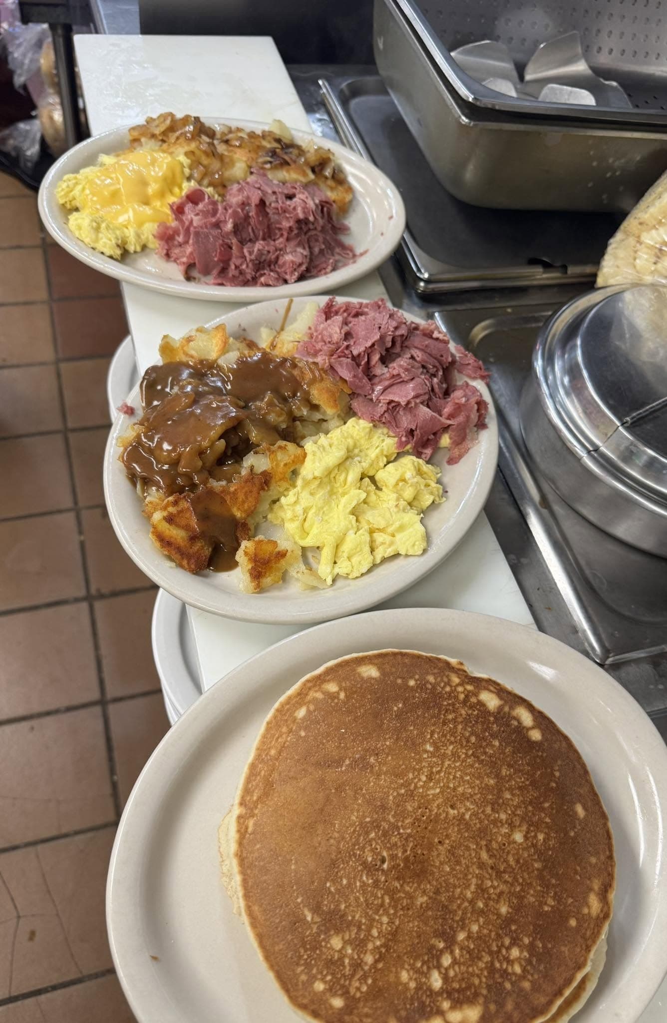 Kitchen pass shot of freshly plated breakfast plates — corned beef, scrambled eggs, hash browns with gravy, and a pancake