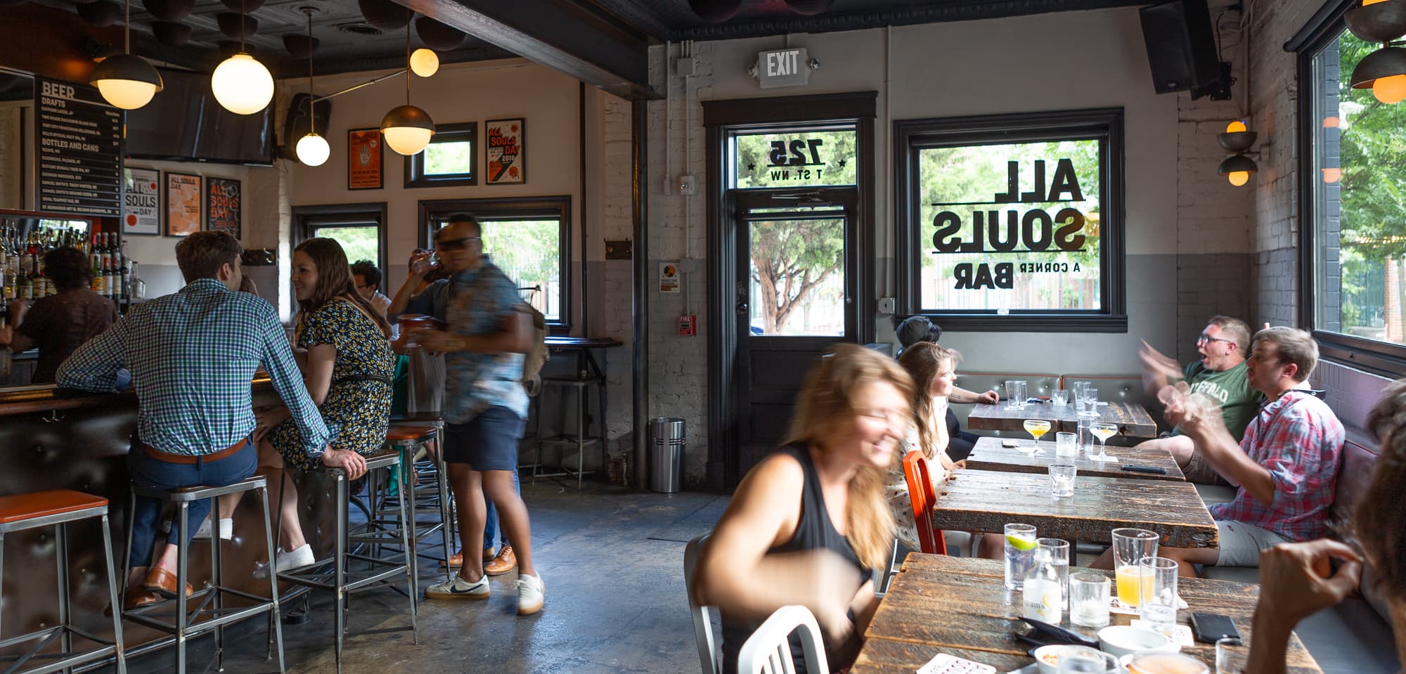 All Souls Bar's warm interior packed with patrons at the bar and communal tables, the reversed logo visible on the front windows