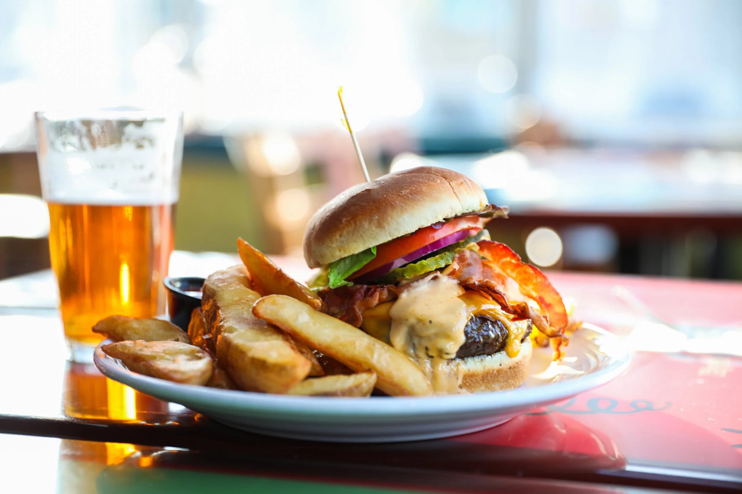 A bacon cheeseburger piled high with fries next to a glass of house draft beer on a sunlit table