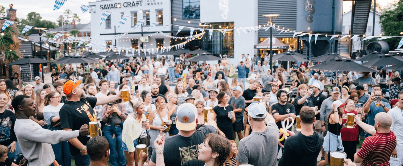 A large crowd raising beers on Savage Craft Ale Works' string-lit outdoor patio, with the brewery's building and Bavarian-flag bunting behind