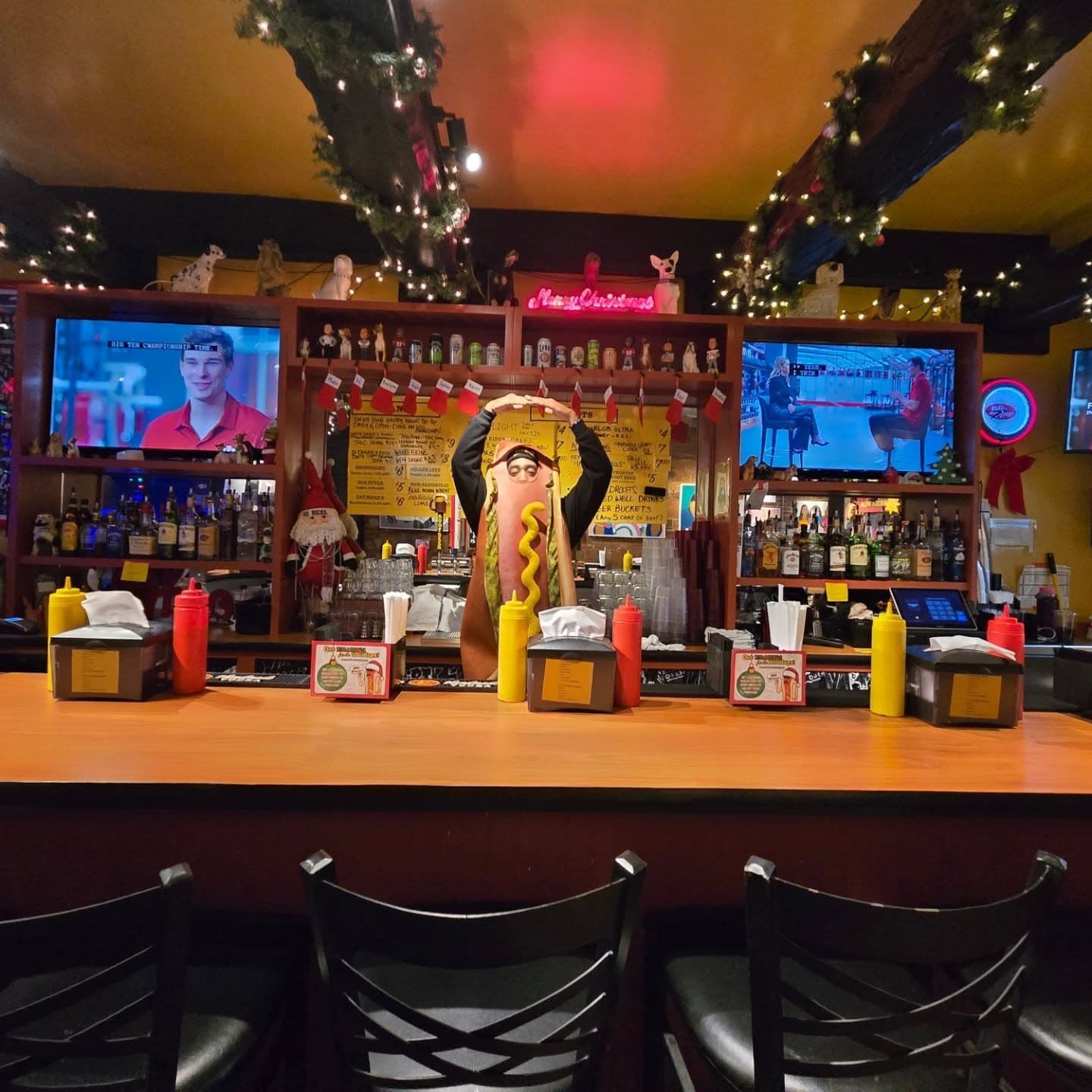 Dirty Frank's Hot Dog Palace bar decked out for the holidays, with a giant hot dog mascot statue behind the counter, condiment bottles lined up, and a Big Ten Championship broadcast on the TVs