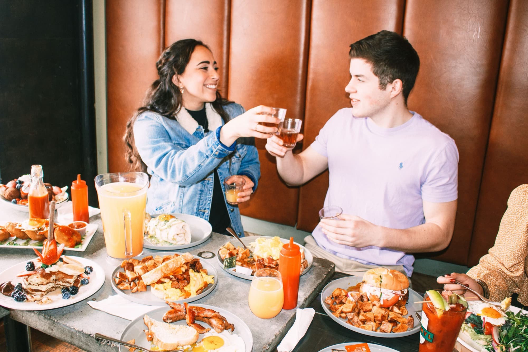 A couple clinking small glasses in a toast over a table loaded with brunch plates, waffle fries, pancakes, and mimosas at Ethyl & Tank