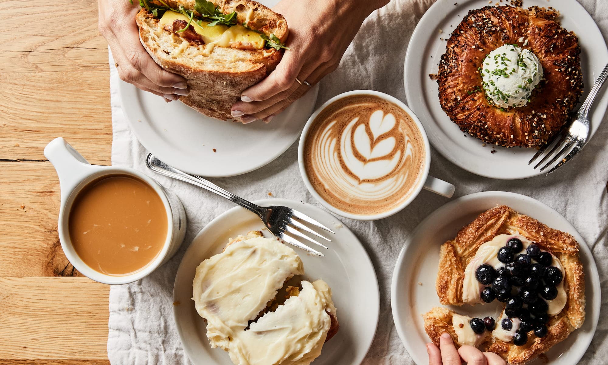A latte, cream cheese frosted cinnamon roll, blueberry pastry, and an everything-seasoned bagel with herbed cream cheese spread on a table at Fox in the Snow