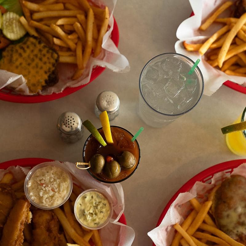 An overhead spread of burger and fried-fish baskets with fries, coleslaw, and tartar sauce alongside a Bloody Mary and a soda at Varsity Club