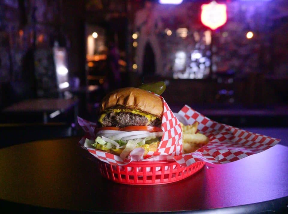 A loaded cheeseburger with a pickle skewer and fries in a red plastic basket on a dark table, neon signs glowing behind the bar