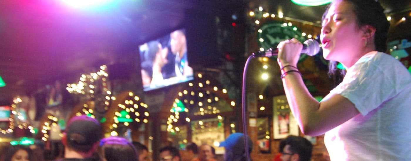 A singer performing karaoke into a microphone at Crunchy's, the bar's string-lit arches and a TV glowing behind the crowd