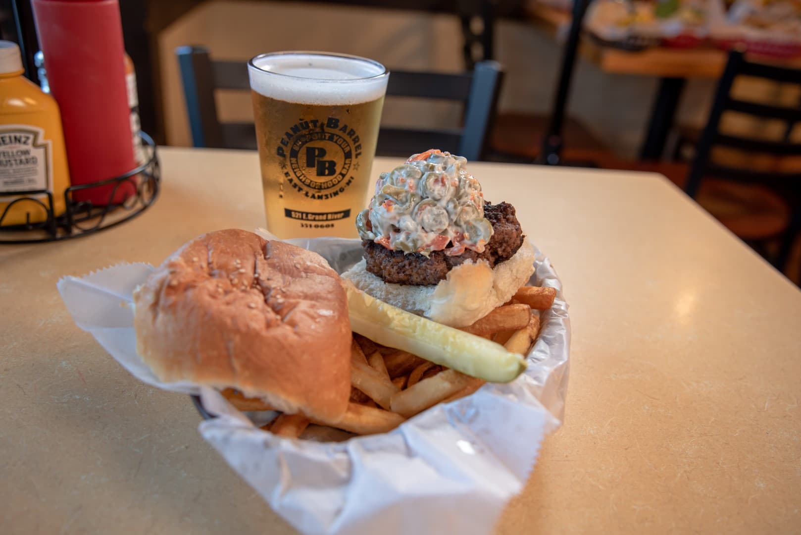A baby burger topped with relish alongside fries and a pickle spear in a basket, next to a Peanut Barrel-branded pint of beer