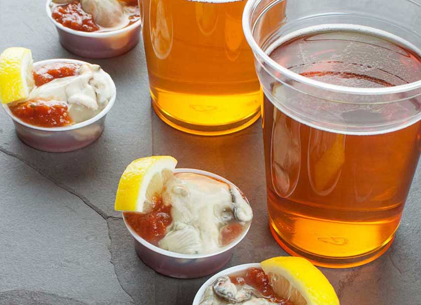 Cups of fresh-shucked oysters on ice with cocktail sauce and lemon next to two glasses of beer