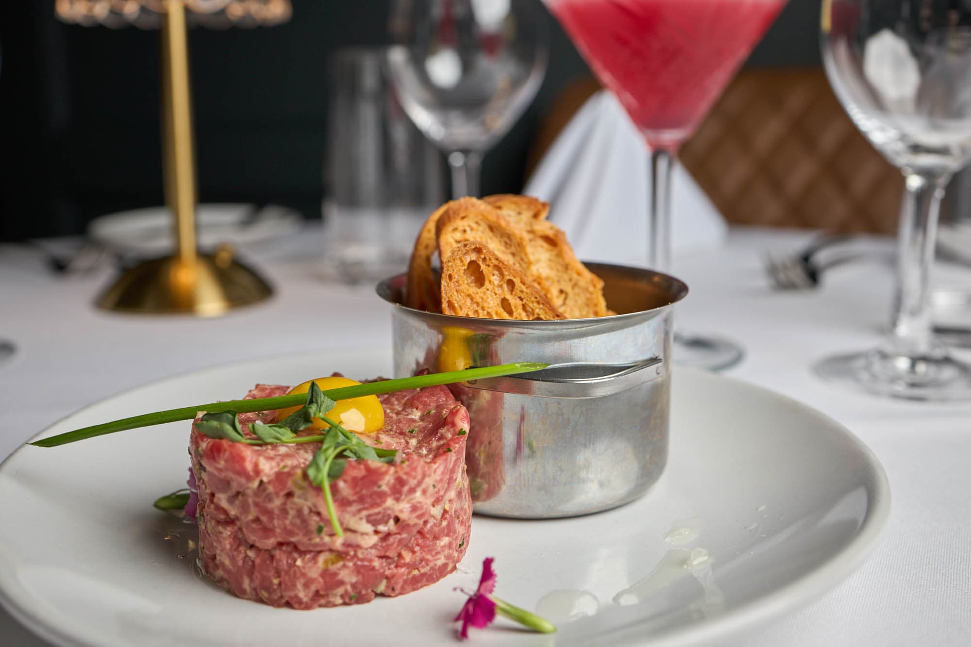 Steak tartare with toasted bread, a martini glass and wine glasses on white tablecloth at Kennedy's Steakhouse