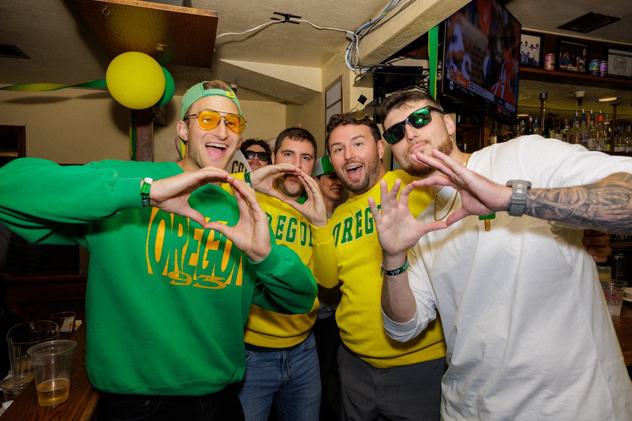 A group of fans in green and yellow Oregon gear throwing the O hand sign at the bar inside Rennie's Landing