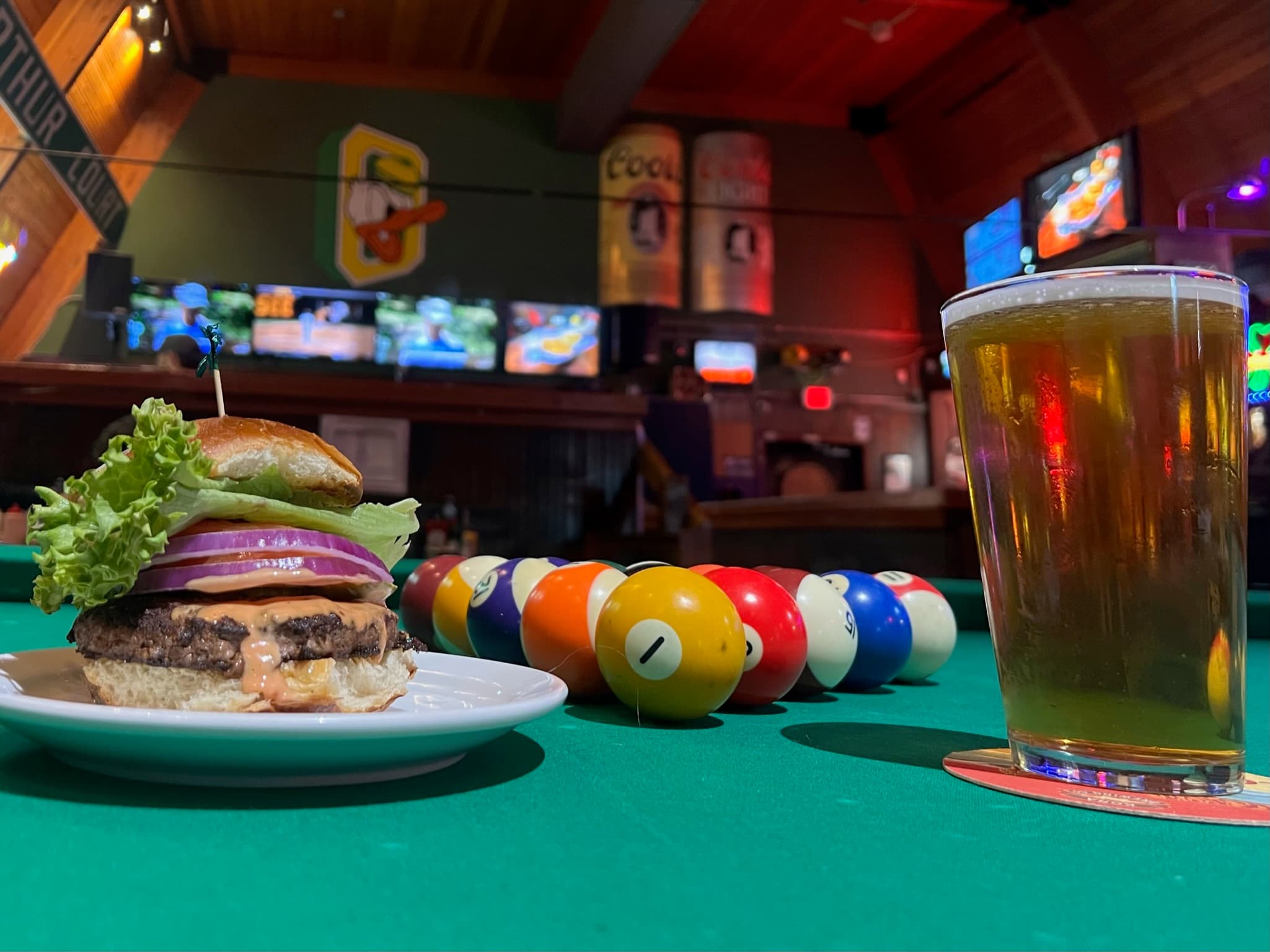 A cheeseburger and a pint of beer set on the felt of a pool table, TVs and Oregon decor glowing in the bar behind