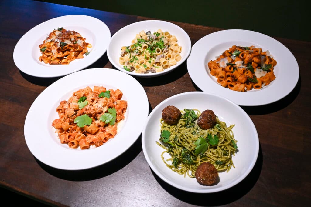 Five plates of pasta on a dark wood table at The Spot — cracker-thin pizza, rigatoni bolognese, and a pesto spaghetti with meatballs