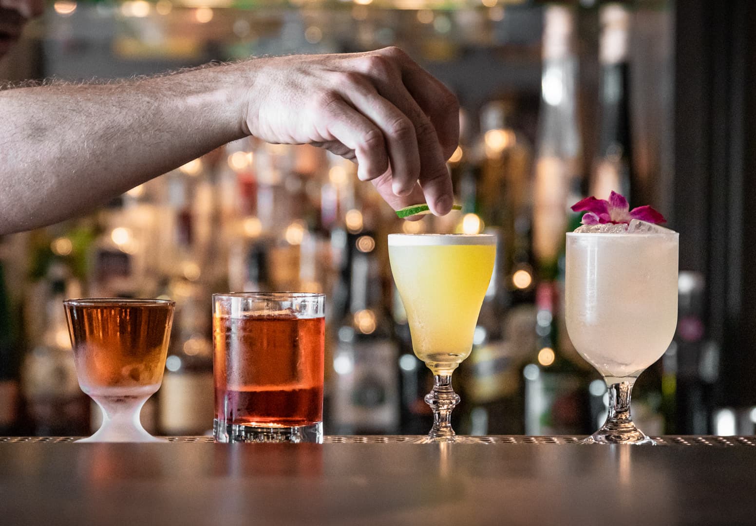 A bartender garnishing a row of four classic cocktails on the bar at Ward Eight, bottles glowing behind on the back bar