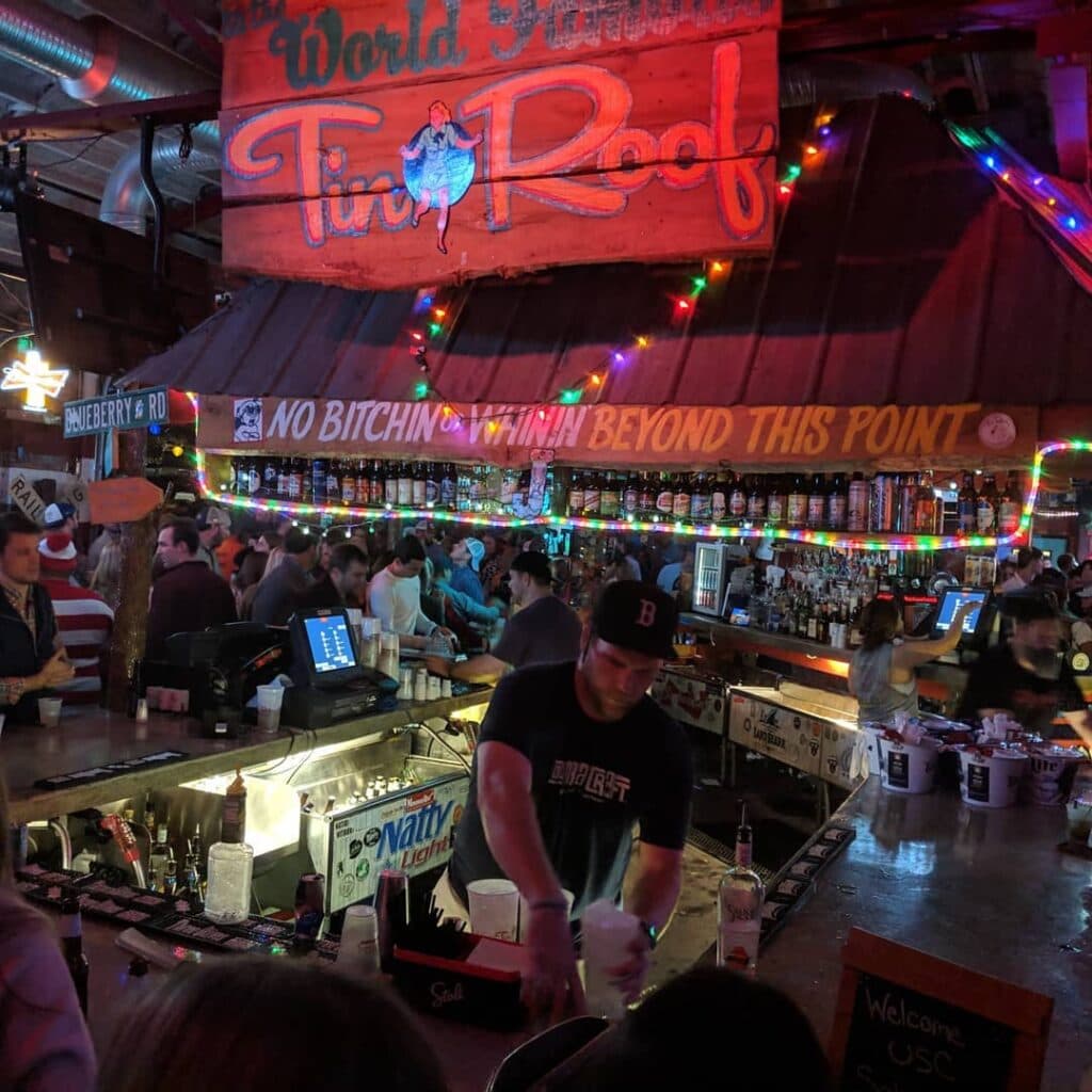 A bartender working the Tin Roof bar under its glowing red neon sign, string lights strung along the ceiling
