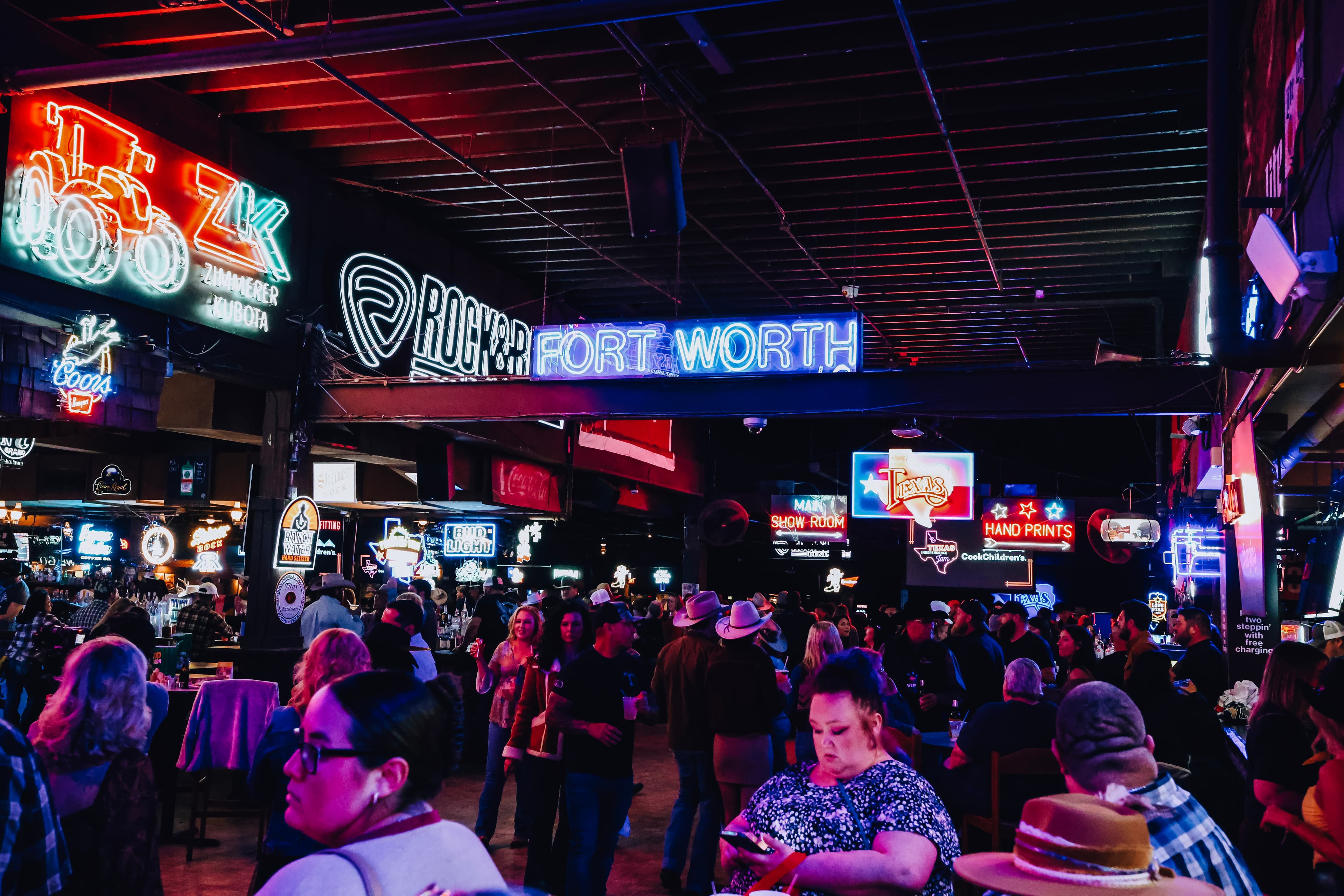 A packed crowd inside Billy Bob's Texas under a wall of neon signs, including a lit-up 'Fort Worth' sign overhead