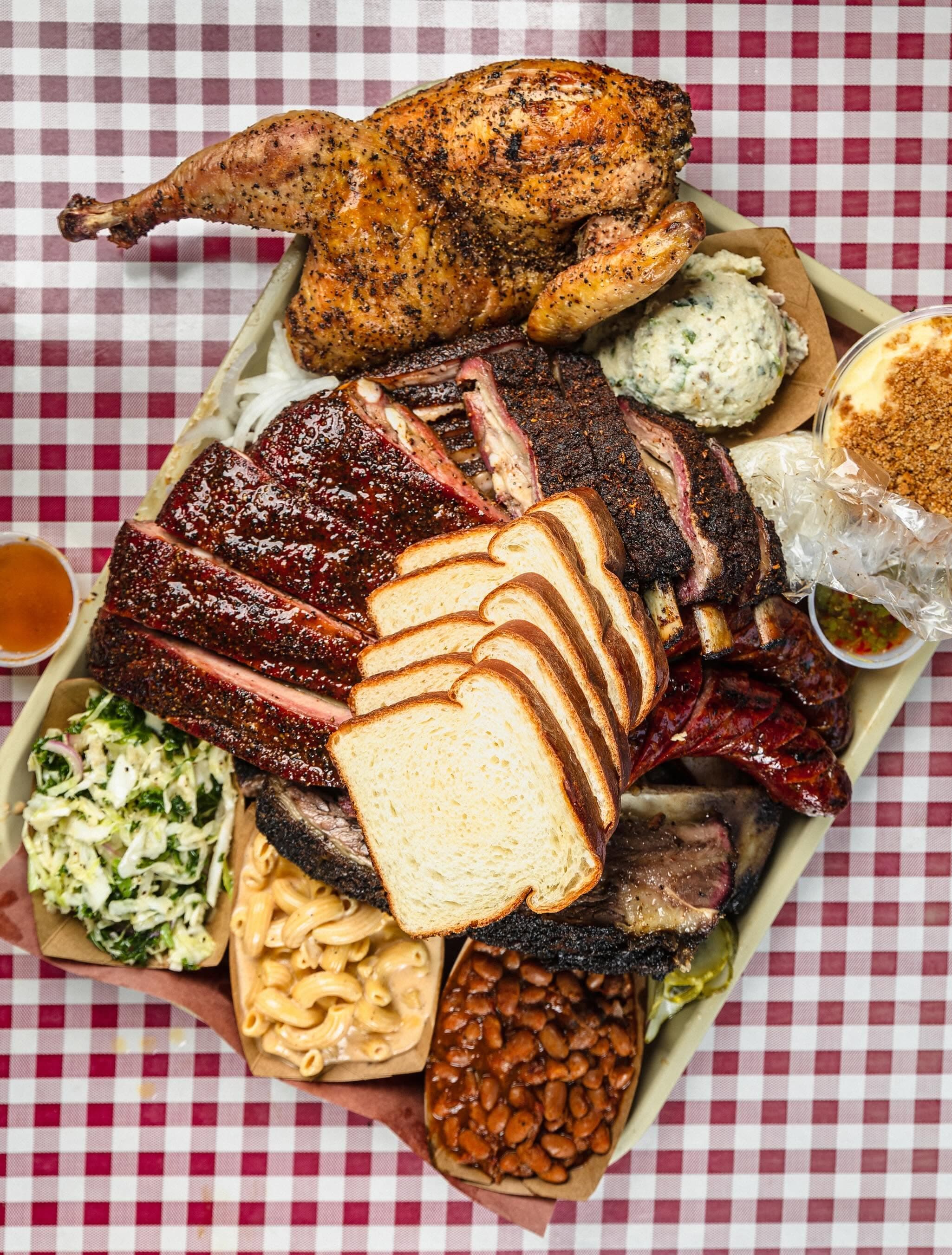 An overhead tray of Goldee's smoked chicken, brisket, ribs, sausage, white bread, and sides on a checkered tablecloth