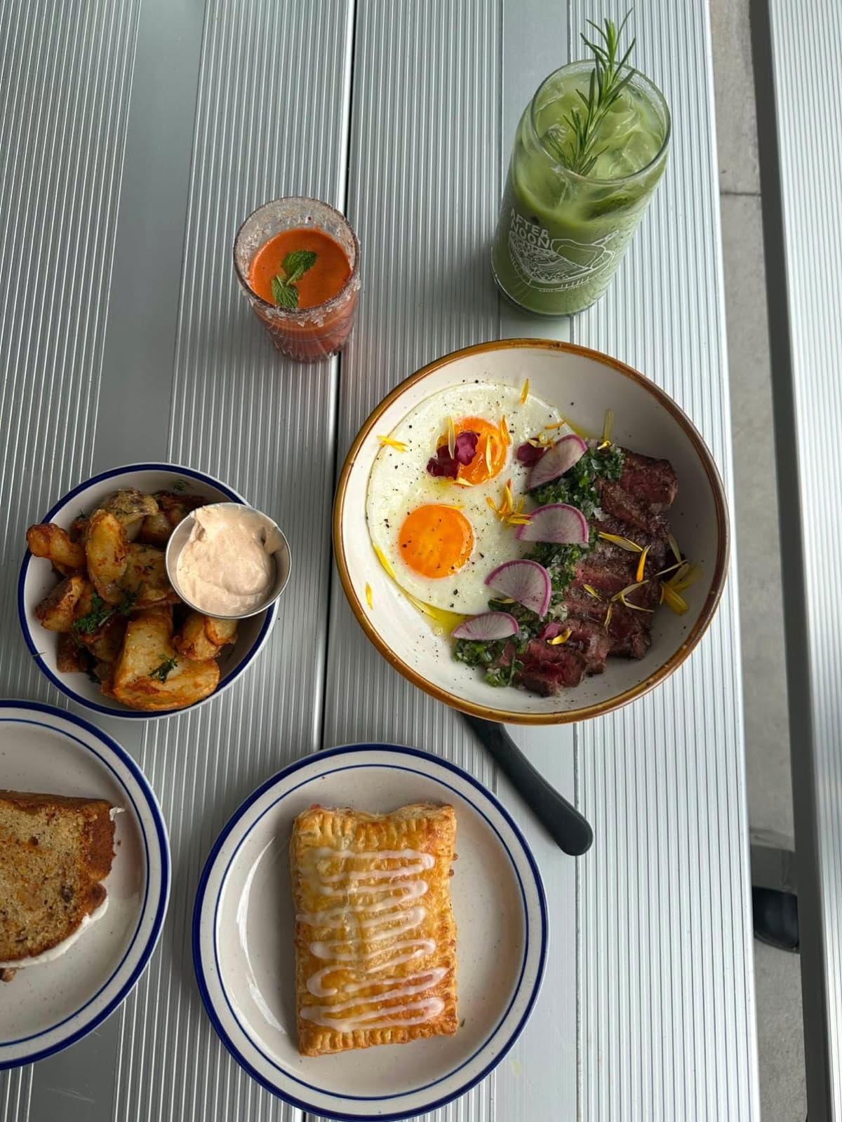 An overhead brunch spread at Afternoon — a steak-and-eggs bowl, crispy potatoes, a pastry, and a branded green juice