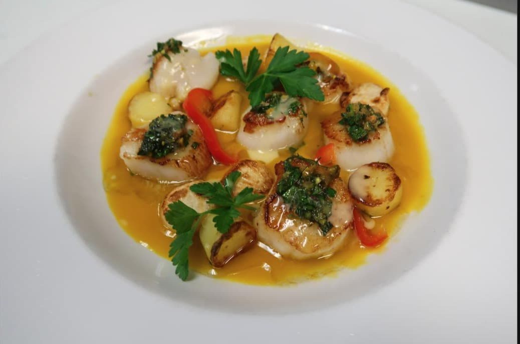 A plate of seared scallops with charred red peppers, roasted potatoes, and herb chimichurri in a bright citrus butter sauce