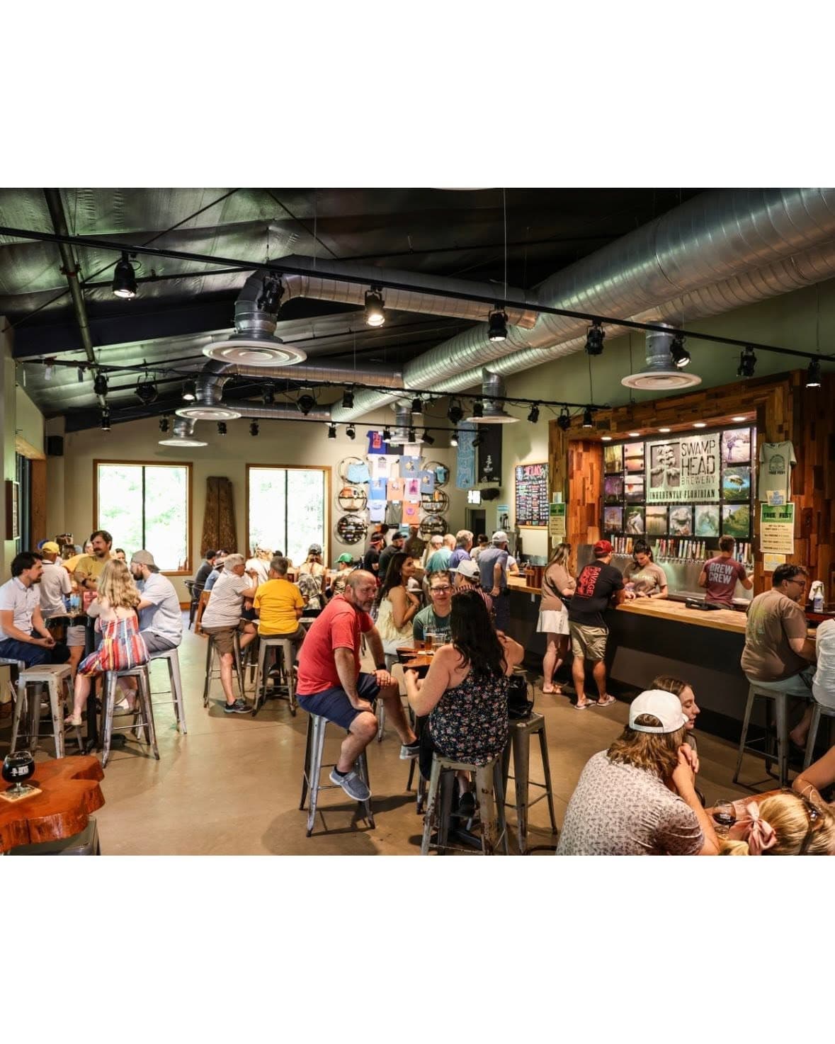 A packed Swamp Head Brewery taproom with people at high-top tables and along the bar under the brand's tap wall