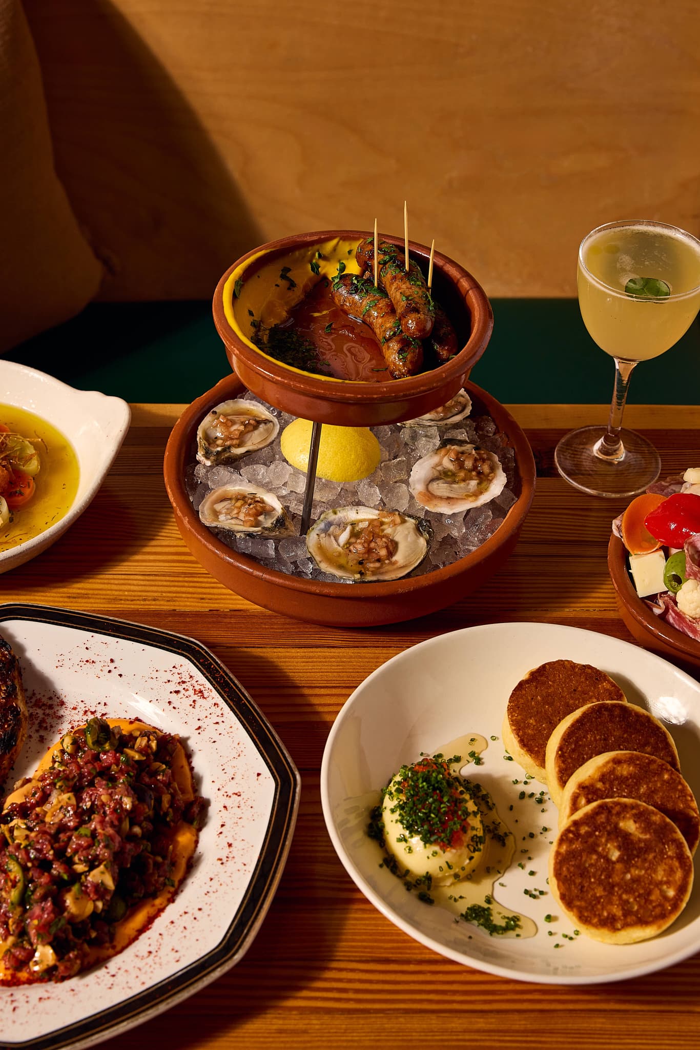 Small-plate spread at Nancy's Hustle — grilled sausages over romesco, chilled oysters on ice, beef tartare, and cornbread cakes with a cocktail