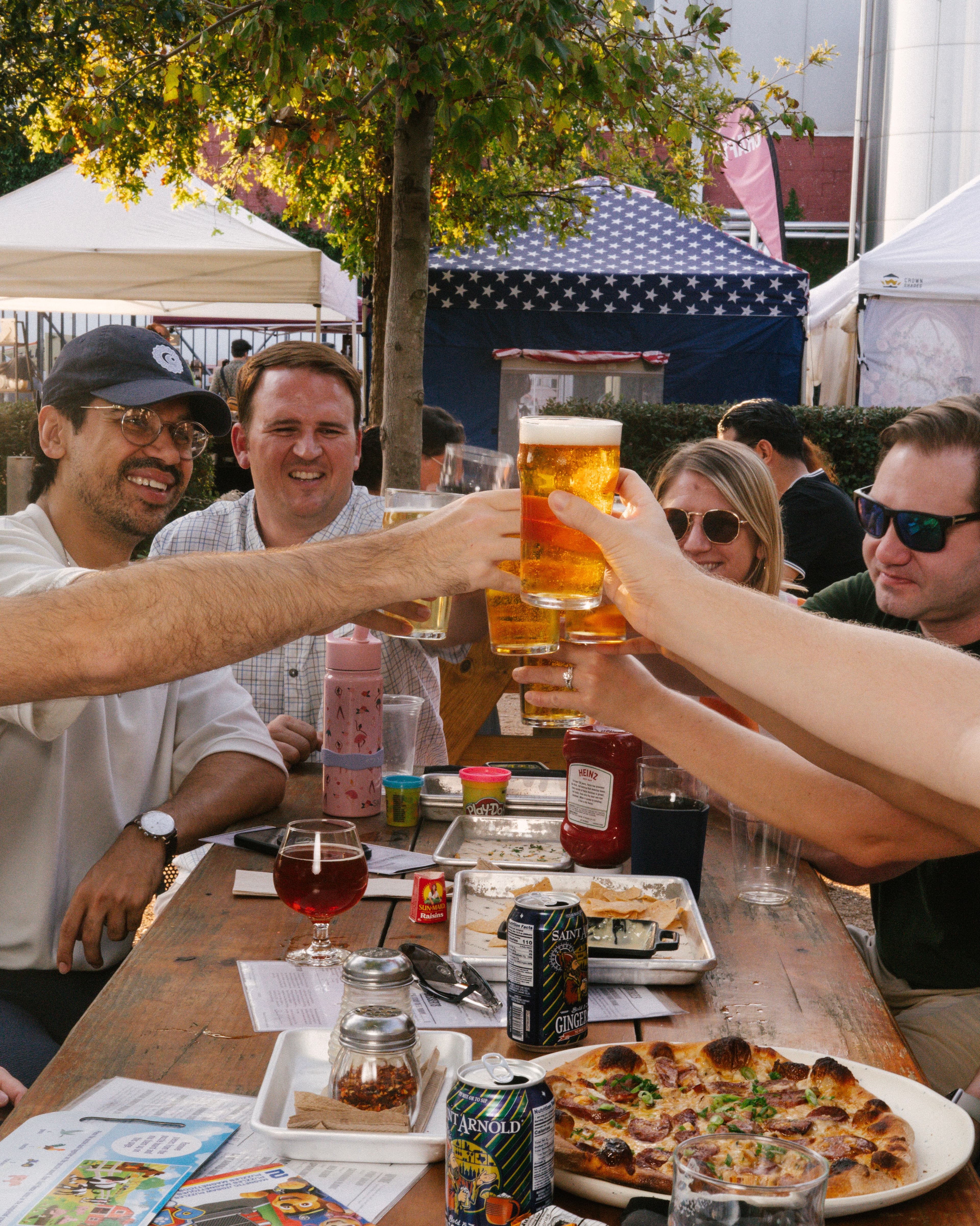 Friends clinking pint glasses of Saint Arnold beer at an outdoor picnic table with pizza and cans of Fancy Lawnmower and Spicy Ginger