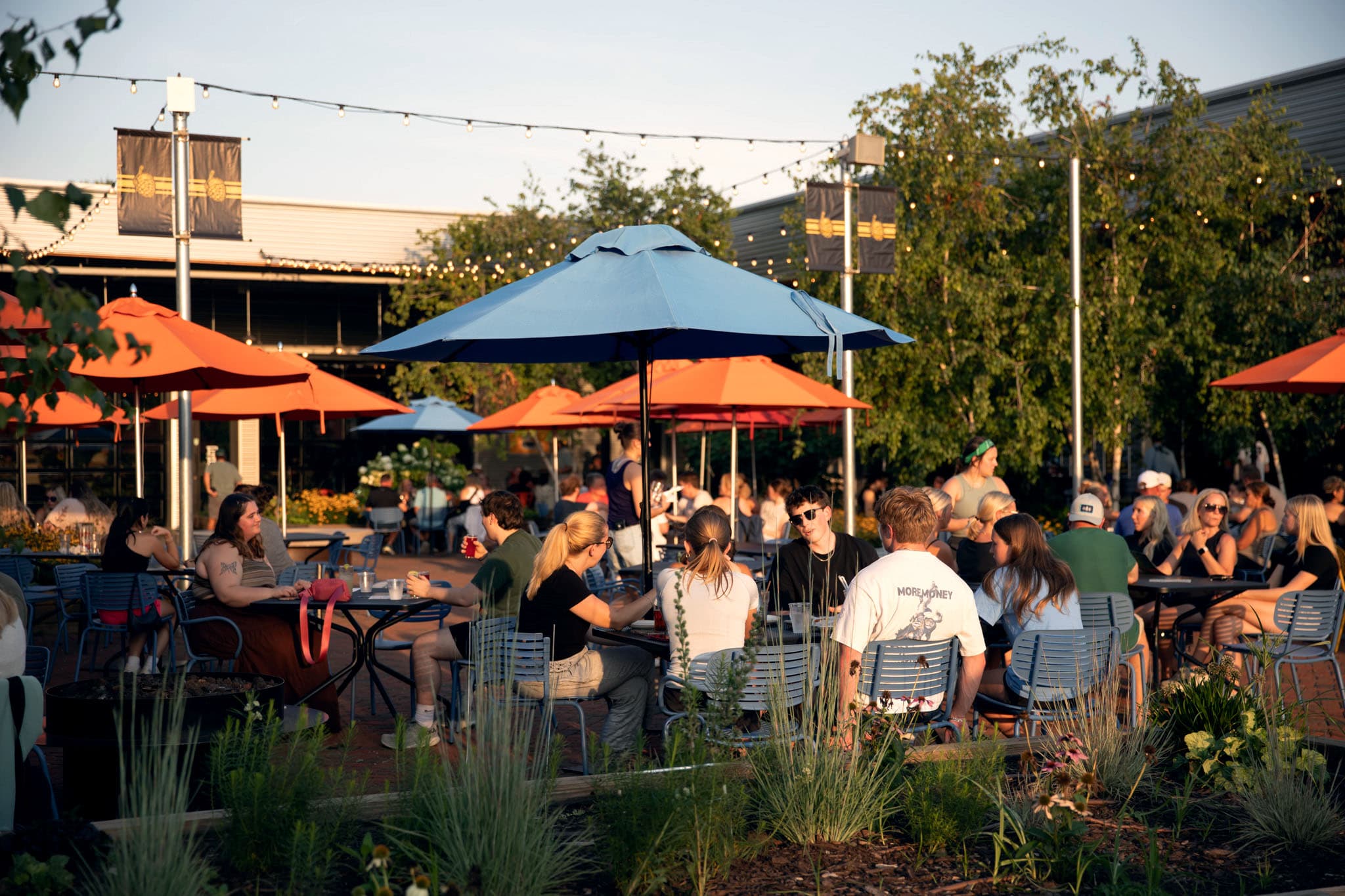 The umbrella-shaded outdoor patio at Big Grove Brewery packed with people at tables around a fire pit at golden hour